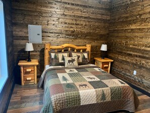 A log cabin-style bedroom with a wooden headboard, two bedside tables with lamps, a quilted bedspread, and a window on the left.