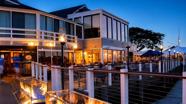 Een restaurant aan het water met terras, een dek met vuurplaats en een modern gebouw met grote ramen.
