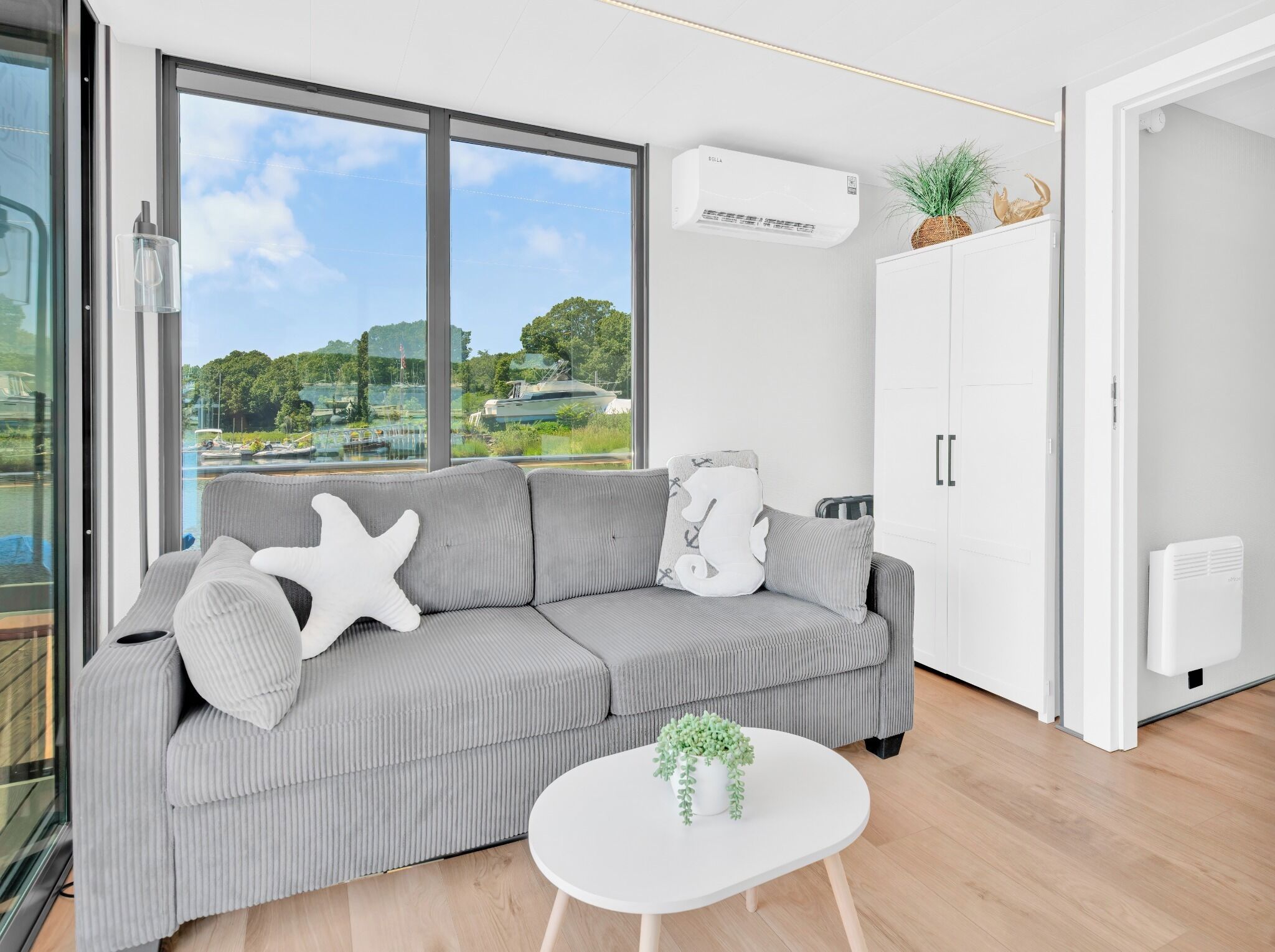 Een moderne woonkamer met een grijze bank, een witte salontafel en uitzicht op een jachthaven door grote ramen.