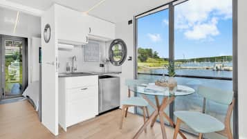 Eine moderne Küche mit weißen Schränken, einer Edelstahl-Spüle und einem Glastisch mit Holzkästen. Ein großes Fenster bietet Blick auf einen Hafen und Grünflächen.