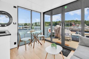 A modern interior with large windows, a round mirror, a table with chairs, and a view of boats and water.