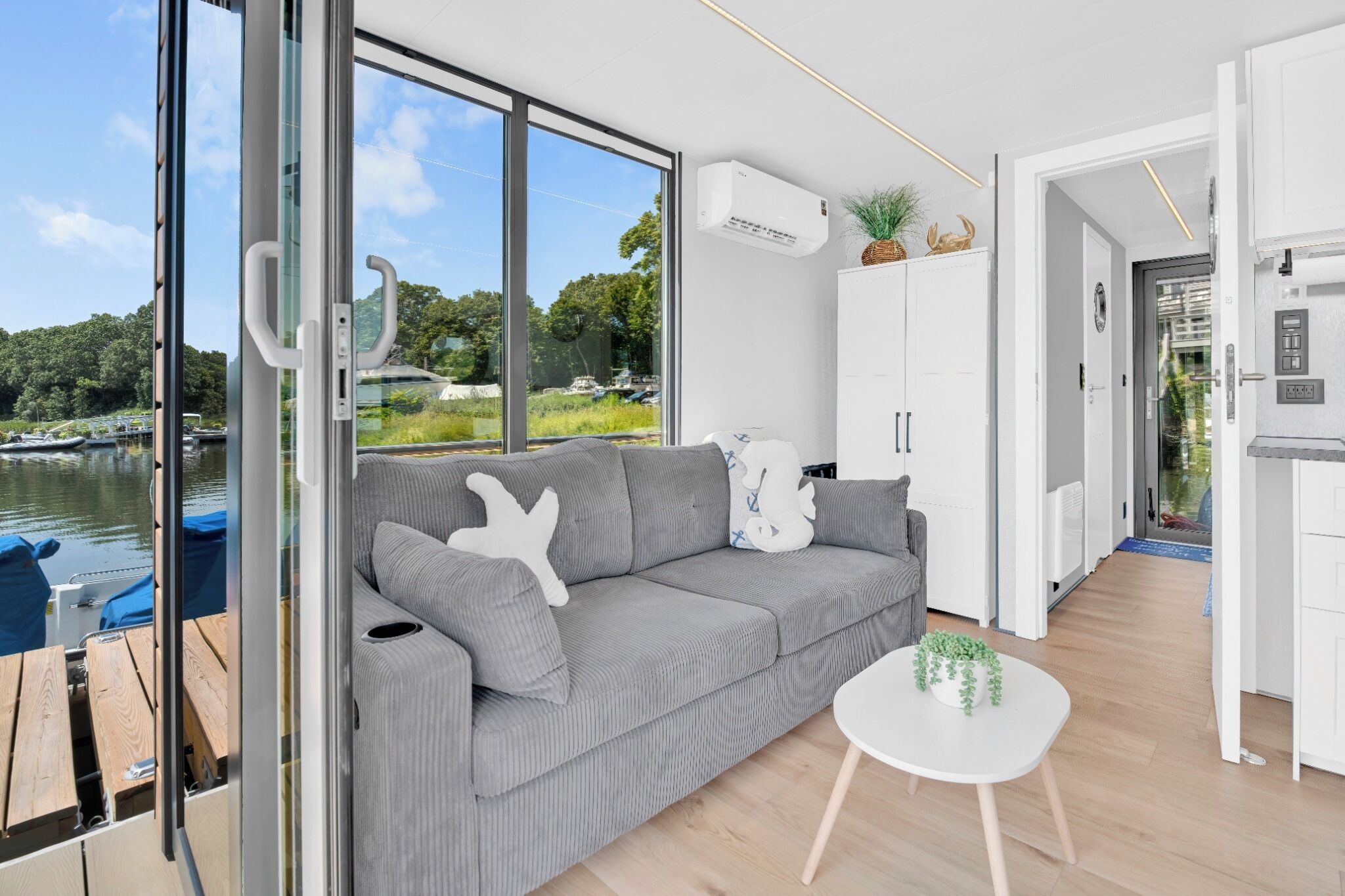 A modern living room with a grey sofa, a small white table with a plant, and a view of a lake with boats.