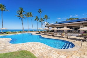 A swimming pool with a curved edge, surrounded by lounge chairs and umbrellas, with palm trees and a view of the ocean.