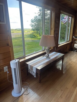 A room with wooden walls, a large window with an American flag, a standing fan, a table with a lamp, and a view of a green landscape.