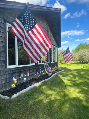 A house with two American flags, a wooden exterior, and a flower bed with various flowers.