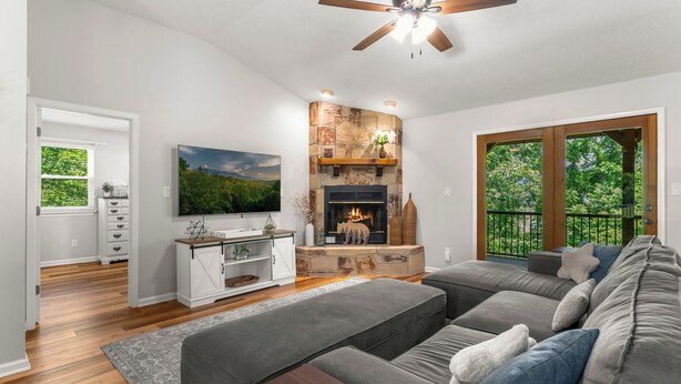A living room with a fireplace, a ceiling fan, and a large window with a view of trees. - Modern Family Cabin in the Smokies + Mtn Views! (Pigeon Forge)