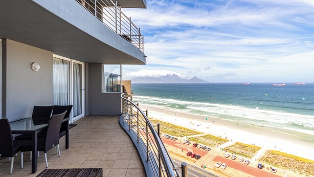 Een balkon met uitzicht op het strand en de oceaan, een eethoek met stoelen en een tafel, en een parkeerplaats met auto's.