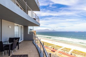 A balcony with a view of the beach and ocean, a dining area with chairs and a table, and a parking lot with cars. - Blu Horizon
Modern Beachfront 3BR with Epic Table Mountain Views (Cape Town)