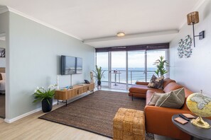 Ein modernes Wohnzimmer mit einer Couch, einem Flachbildfernseher, einem Couchtisch und Meerblick durch große Fenster.