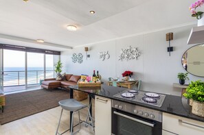 Eine moderne Küche mit einer großen Kochinsel, ein Essbereich mit Aussicht und ein Wohnbereich mit Sofa und Pflanze.