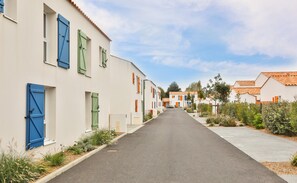 Eine schmale Straße gesäumt von weißen Gebäuden mit blauen und grünen Fensterläden, die zu einem Wohngebiet mit Bäumen und Sträuchern führt.