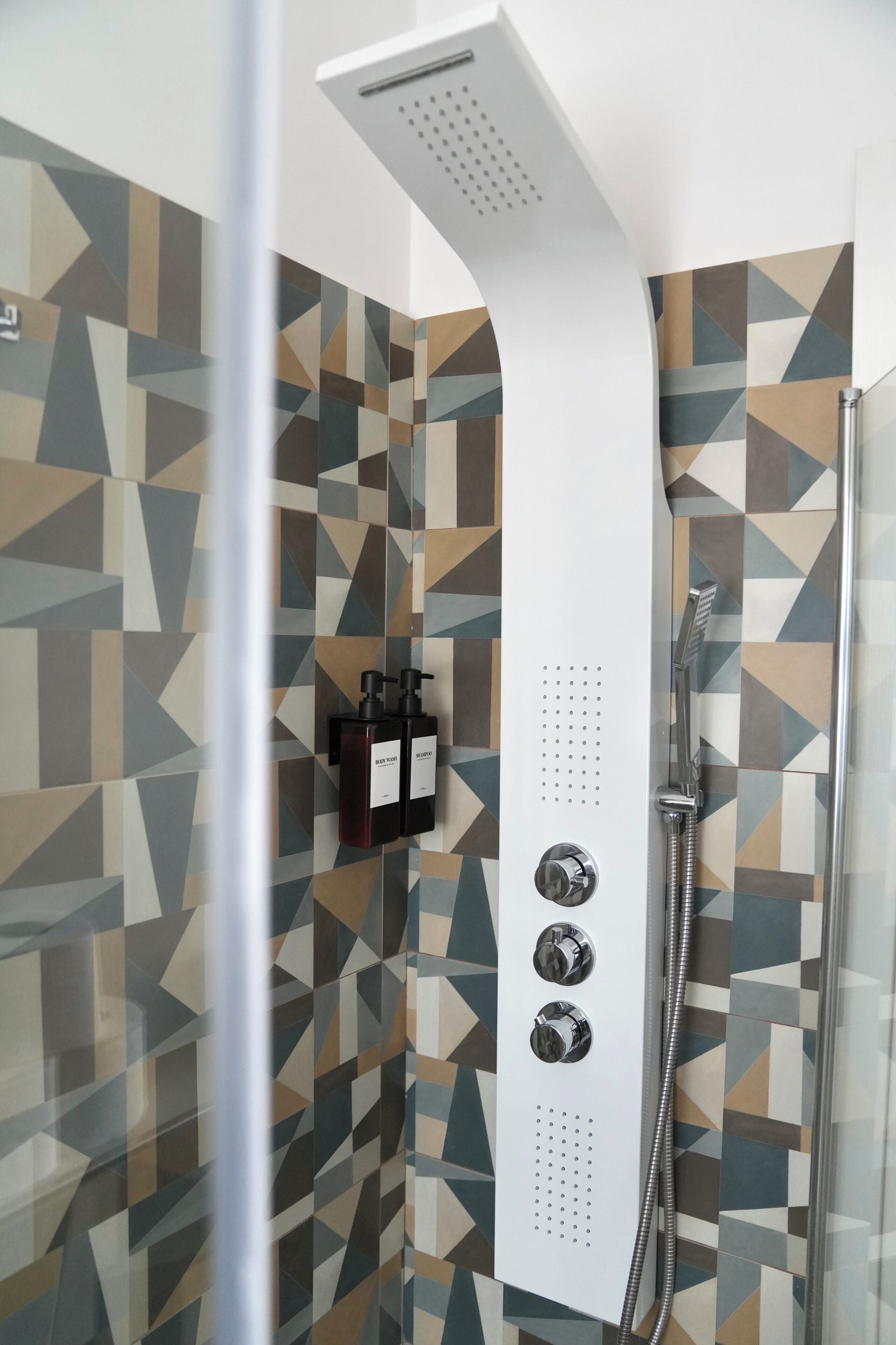 Une douche avec un carrelage aux motifs géométriques, un pommeau de douche à main et deux bouteilles de shampoing.