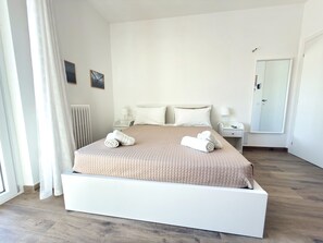 Ett modernt hotellrum med en stor säng, vita sängkläder, ett sängbord med en lampa, ett litet bord, en spegel och utsikt över omgivningarna.