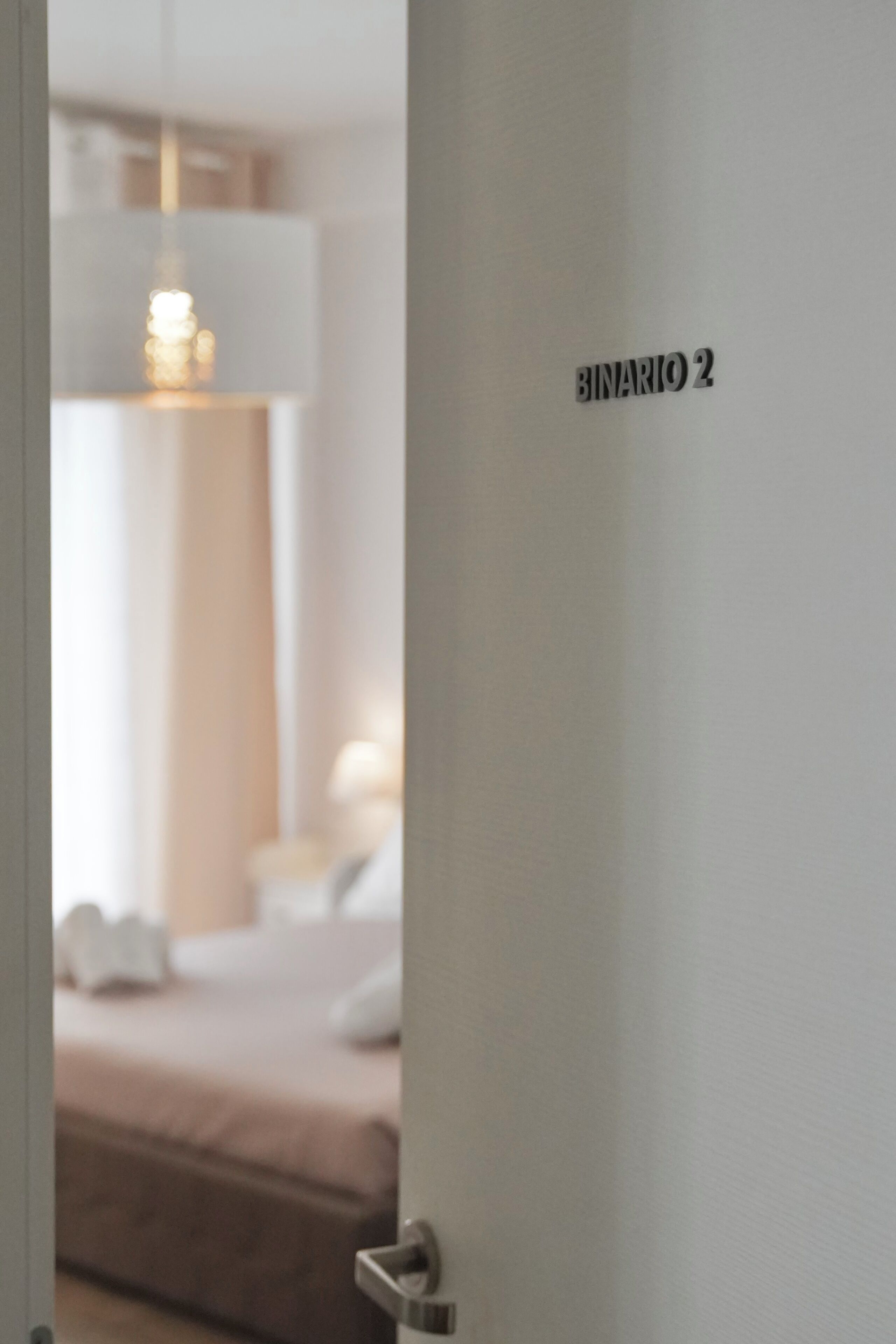 A door with a sign reading 'BINARIO 2' and a blurred interior view of a room with a bed and a hanging light fixture.