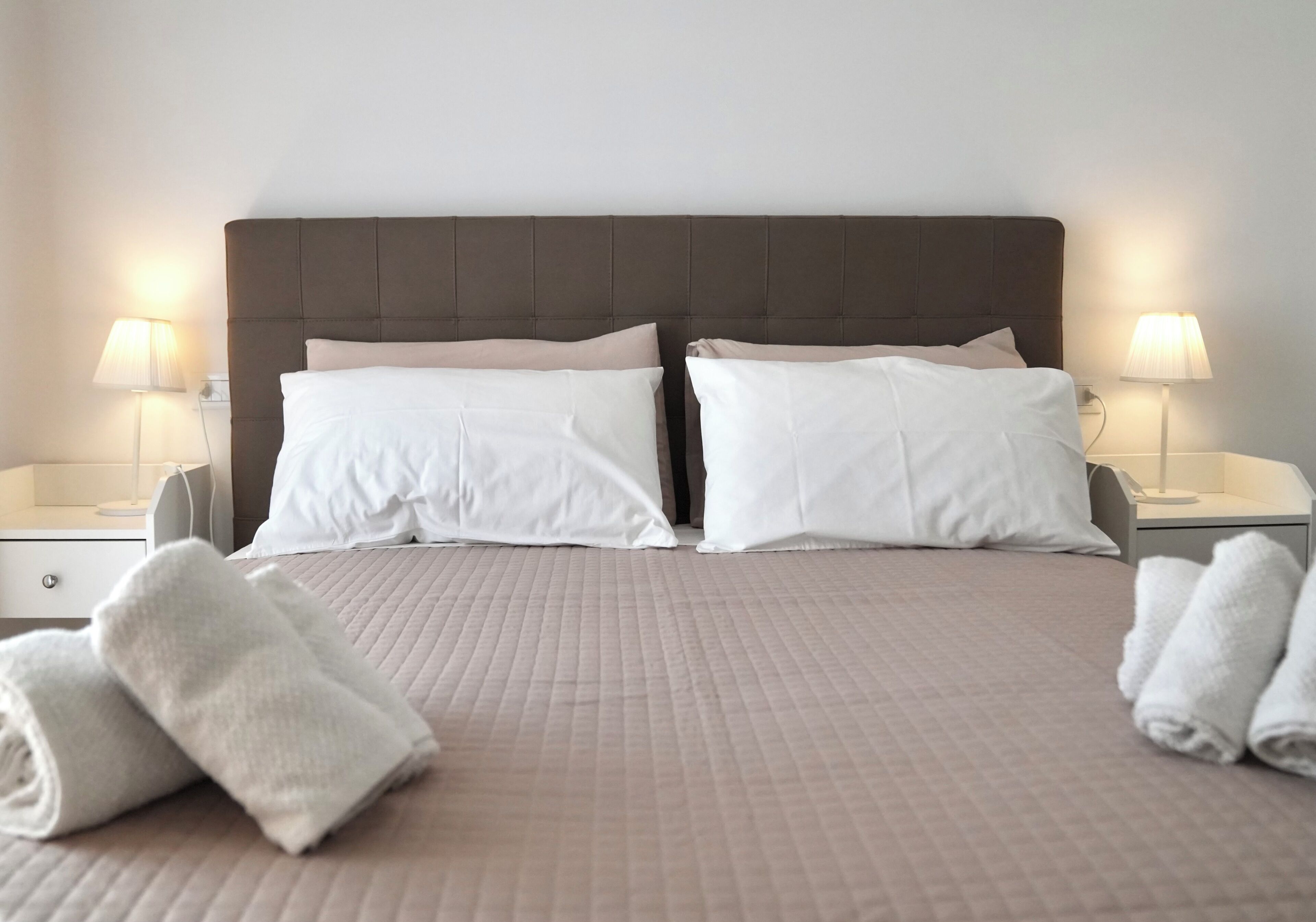 A double bed with a quilted headboard, two bedside lamps, and rolled towels on the bed.