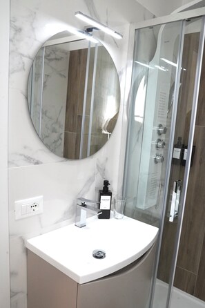 A modern bathroom with a marble wall, a round mirror, a glass shower enclosure, and a white sink. - Termoli Station (Termoli)