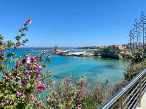 A coastal view with clear blue waters, rocky cliffs, and buildings in the background. - Katy's Holiday House, Private Villa (Sicilia)