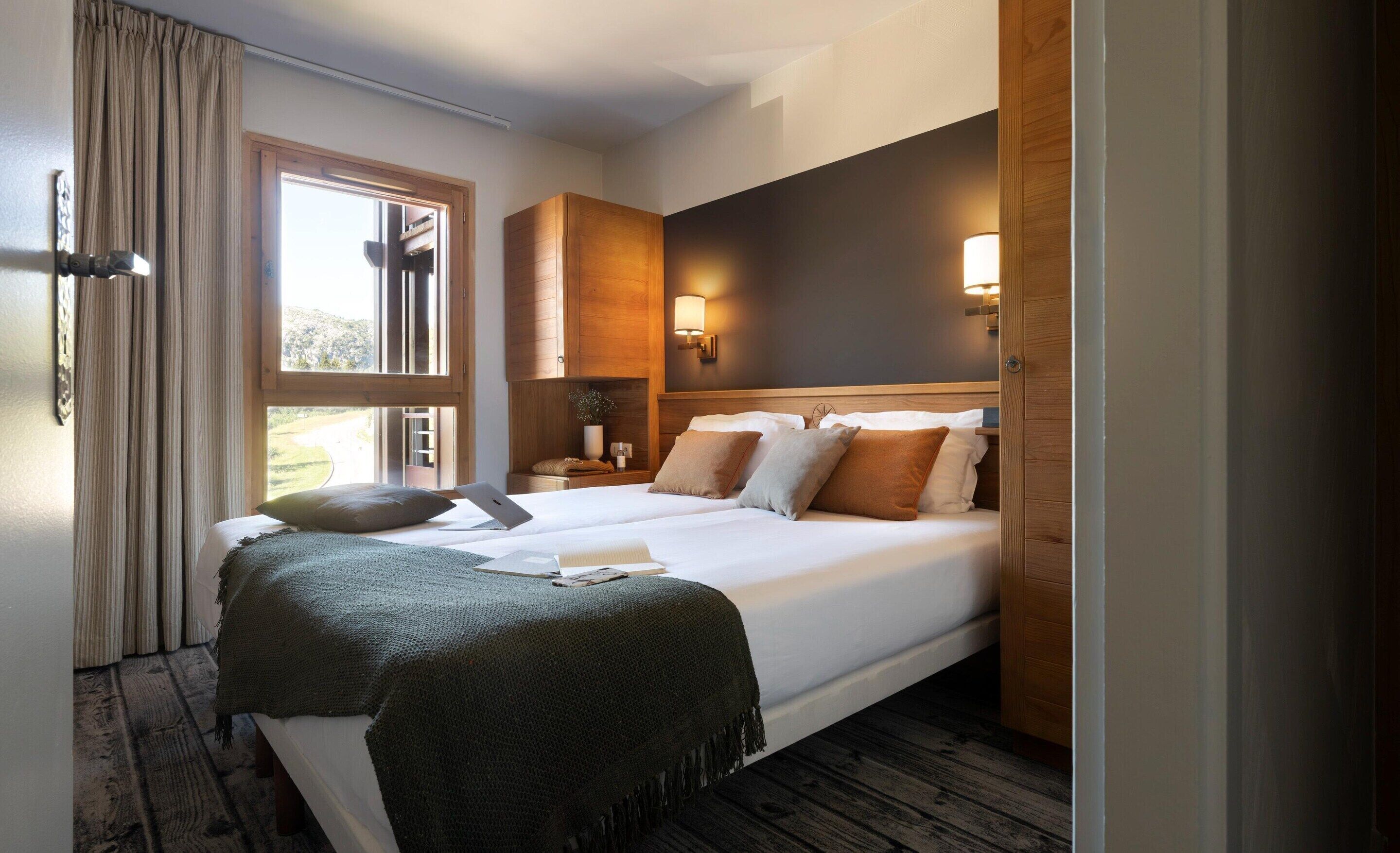 A double bed with a green blanket, two bedside tables with lamps, a window with curtains, and a wooden headboard.