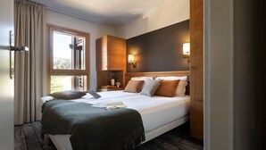 Una cama doble con una manta verde, dos mesitas de noche con lámparas, una ventana con cortinas y un cabecero de madera.