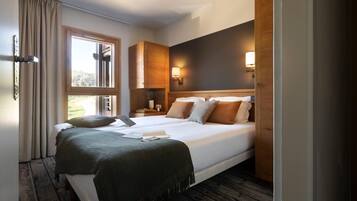 A double bed with a green blanket, two bedside tables with lamps, a window with curtains, and a wooden headboard.