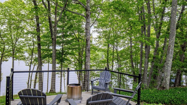 Um terraço com duas cadeiras Adirondack, um braseiro de metal e vista para um lago rodeado por árvores.