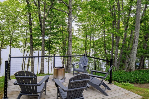 A deck with two Adirondack chairs, a metal fire pit, and a view of a lake surrounded by trees.