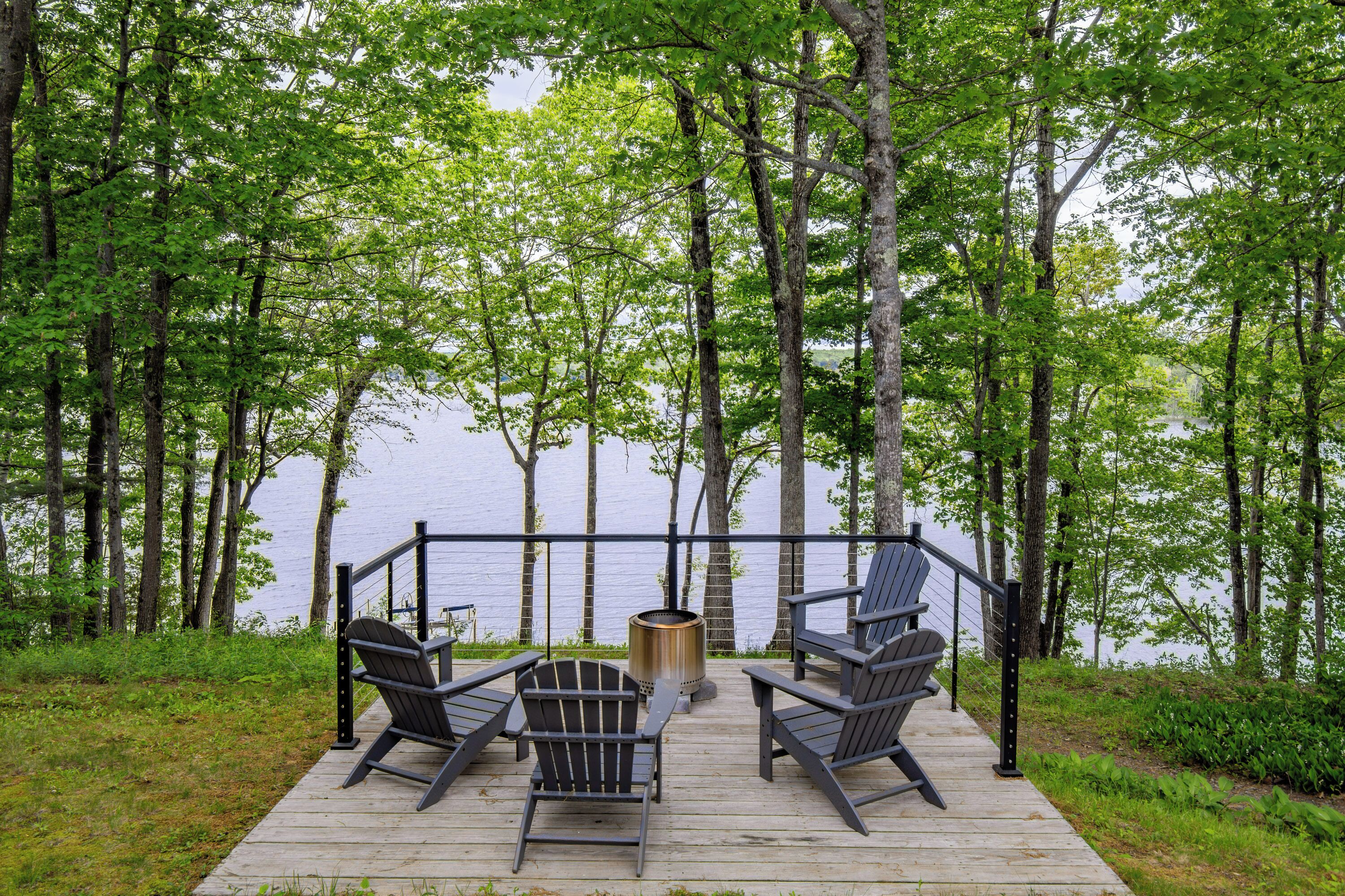 Una terraza de madera con sillas Adirondack y un pozo de fuego con vistas a un lago.