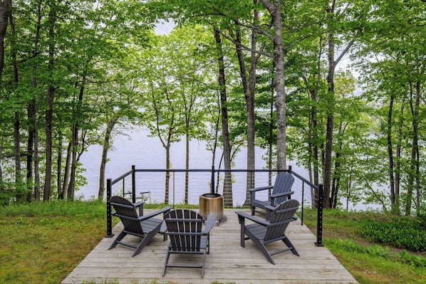 A wooden deck with Adirondack chairs and a fire pit overlooking a lake.