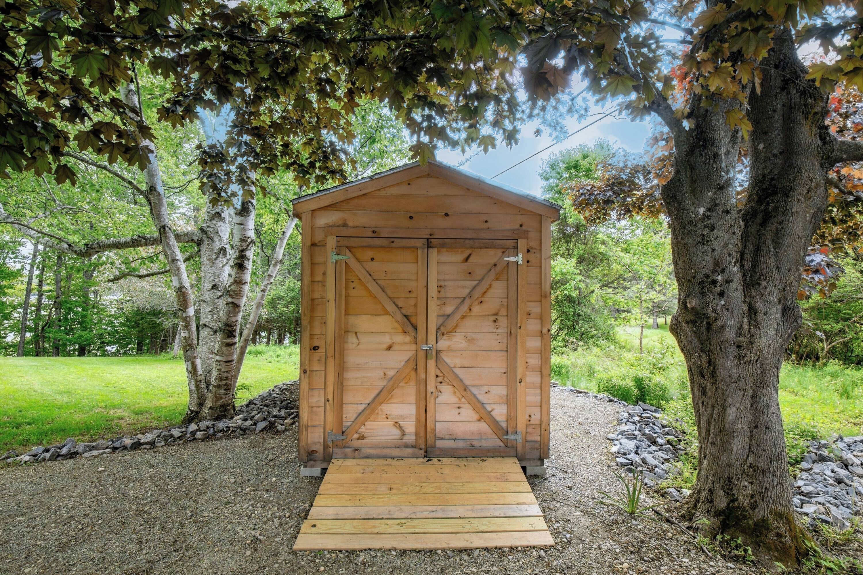 A wooden shed with a cross-braced door, surrounded by trees and gravel.