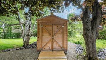 A wooden shed with a cross-braced door, surrounded by trees and gravel.