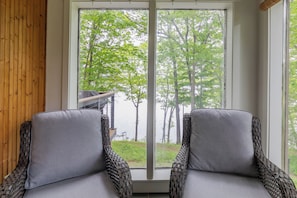 A room with two wicker chairs facing a large window with a view of trees and a dock.