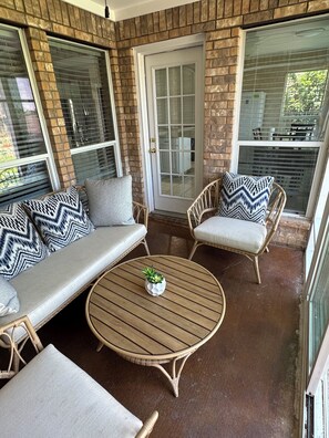 Een overdekt terras met rieten meubilair, een ronde tafel met een plant en een deur met een ruit.