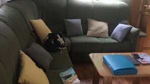 En grøn hjørnesofa med mange puder, et træbord med en blå mappe, og et vindue med udsigt til grønne områder.
