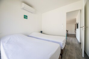 Habitación con dos camas, aire acondicionado instalado en la pared, una mesita y una puerta que da a un pasillo.