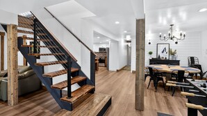 A modern interior with a wooden staircase, a dining area with a large table, and a kitchen in the background.
