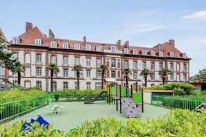 Un patio de juegos con toboganes y estructuras para escalar frente a un edificio histórico grande con muchas ventanas y techo de tejas rojas.