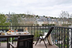 Un balcón con una mesa lista para comer, con vistas a un paisaje urbano que incluye edificios y una aguja de iglesia.