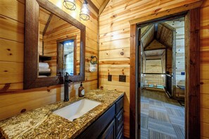 Ein Badezimmer einer Holzhütte mit Waschbecken auf einer Granitarbeitsplatte, einem großen Spiegel und einer Tür zu einem Schlafboden.