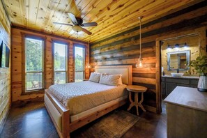 Un dormitorio de cabaña de madera con cama, una mesita redonda pequeña, un ventilador de techo y un baño con lavamanos y espejo.