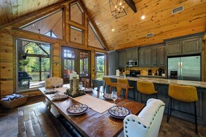 A wooden cabin interior with a dining area, kitchen, and large windows. - Waterfront Retreat + Stocked Fishing Pond! Hot Tub • Firepit • Sleeps 10 (Broken Bow)