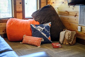 Ein gemütliches Zimmer mit einem großen orangefarbenen Sitzsack, einem dunkelbraunen Pouf und einem gemusterten Kissen. Ein gewebter Korb und eine Schachtel mit Yahtzee-Würfeln stehen auf dem Boden.
