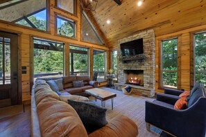 A cozy cabin interior with a stone fireplace, leather sofas, and large windows. - Waterfront Retreat + Stocked Fishing Pond! Hot Tub • Firepit • Sleeps 10 (Broken Bow)