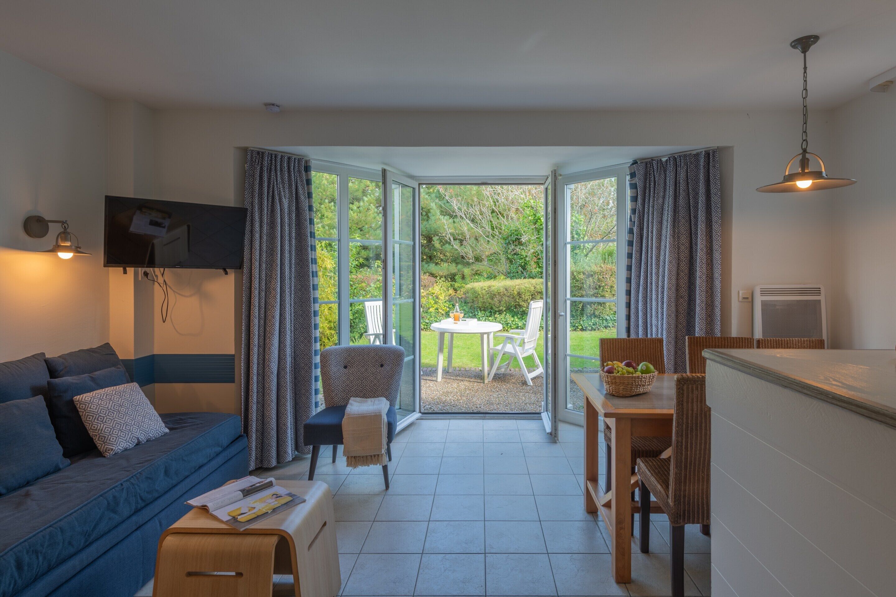 Ein Wohnzimmer mit Sofa, einem kleinen Tisch, einem Sessel und einem Fernseher. Es gibt einen Essbereich mit Tisch und Stühlen sowie einen Blick ins Freie durch große Fenster.