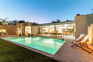 Un área de piscina moderna al aire libre con una terraza de madera, sillas de descanso y un espacio interior bien iluminado.