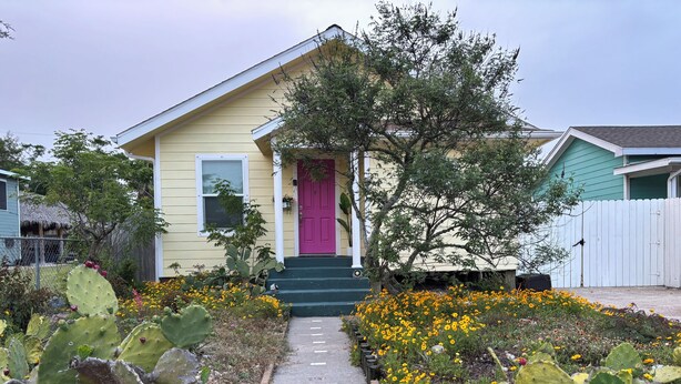 Una casa con puerta rosa, un porche delantero y un jardín con cactus y flores amarillas.