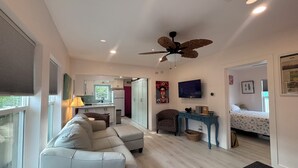 Una sala de estar moderna con un sofá, un ventilador de techo, un televisor y una cocina al fondo.