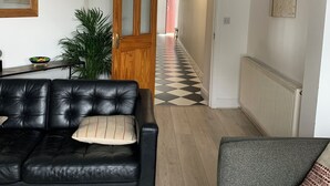Ein Wohnzimmer mit einem schwarzen Ledersofa, einem grauen Sessel, einer Holztür, einem Flur mit Schachbrettfußboden und einem Heizkörper.