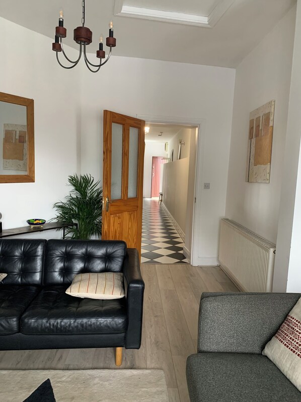 A living room with a black leather sofa, a grey armchair, a wooden door, a hallway with a checkered floor, and a radiator.