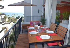 En balkon med et træbord dækket til spisning, en potteplante og udsigt til havet.