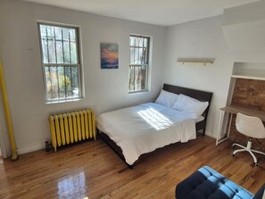 Ein Schlafzimmer mit einem Bett, zwei Fenstern, einer Heizung, einem Schreibtisch mit Stuhl und einem Bild an der Wand.